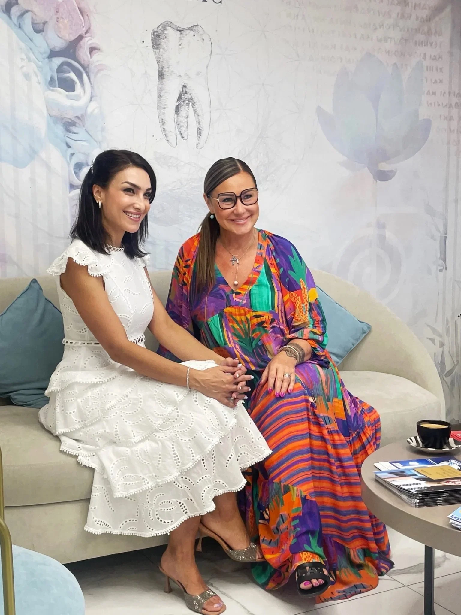 Two women sitting on a cream-colored couch in a room with artistic wall decor. The woman on the left has dark hair, is smiling, and wearing a white dress with eyelet lace. The woman on the right has light brown hair pulled back, wears glasses, a colorful patterned dress, and gold high heels, and is holding hands with the woman on the left. They are sitting close together and smiling.