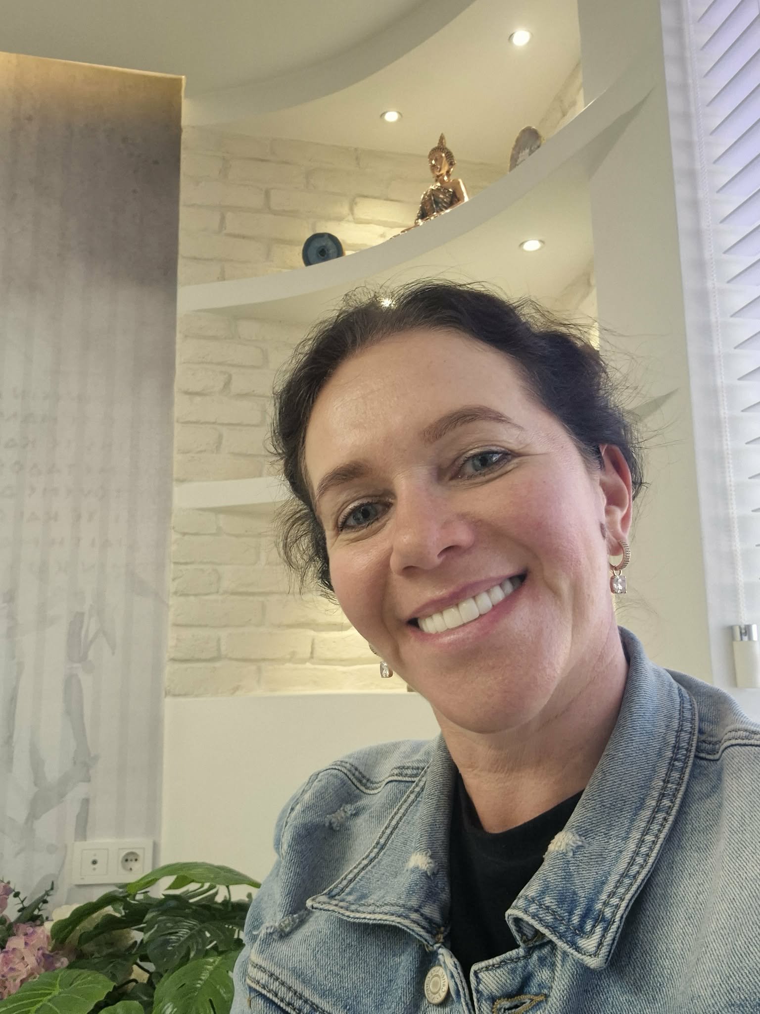 Close-up of a smiling woman with short dark curly hair, wearing earrings and a denim jacket, sitting indoors with a brick wall and white shelves in the background.