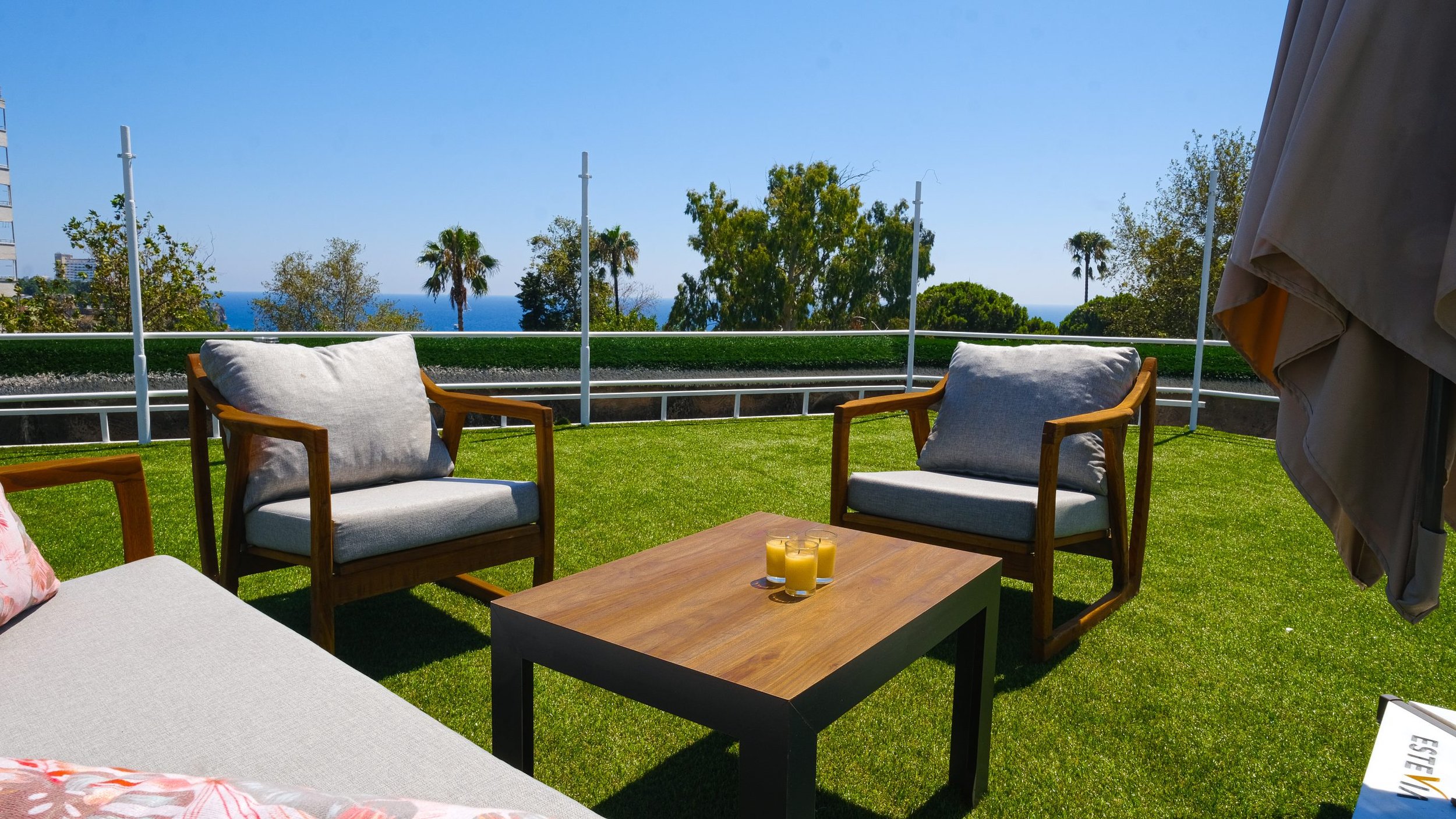 Outdoor patio with wooden chairs, a table with three yellow candles, green artificial grass floor, and a view of trees, palm trees, and the ocean in the distance under a blue sky.